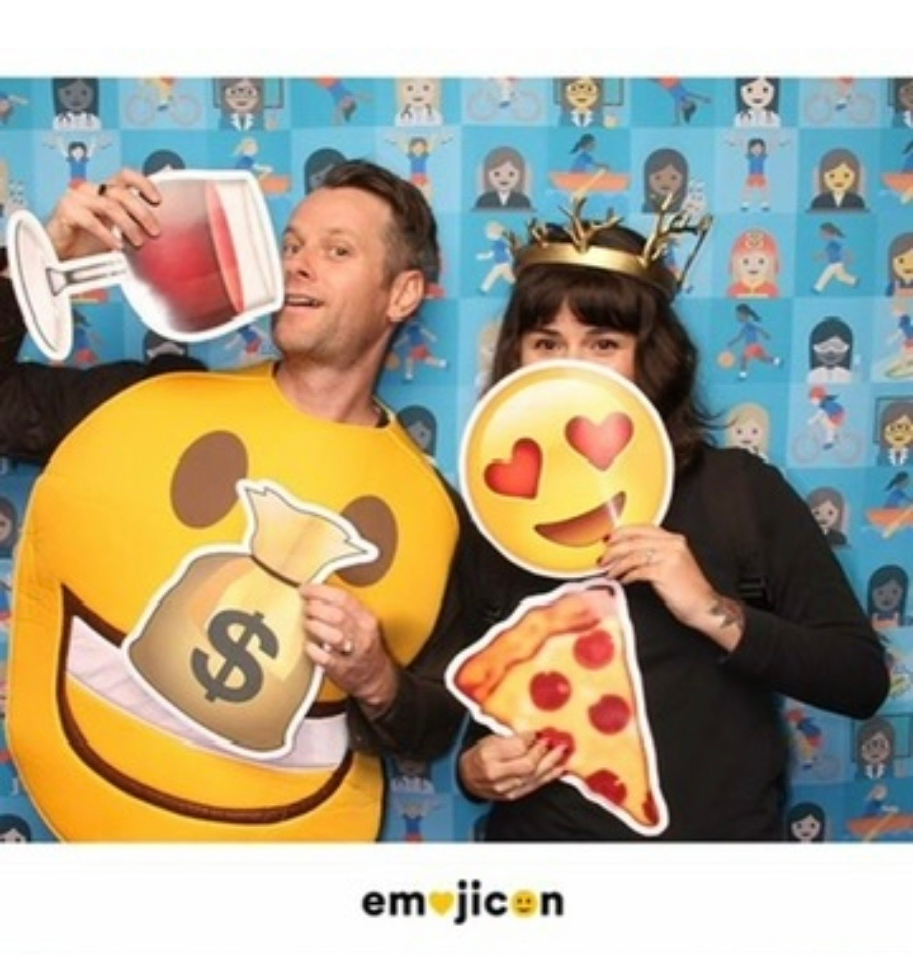 My husband and I, a man and woman in front of an emoji background, pose with emoji props at Emojicon 2016. I am holding a heart-eyes emoji over my face with one hand, and a pizza emoji in the other. Adam is holding a wine emoji and wearing a smiley face emoji.